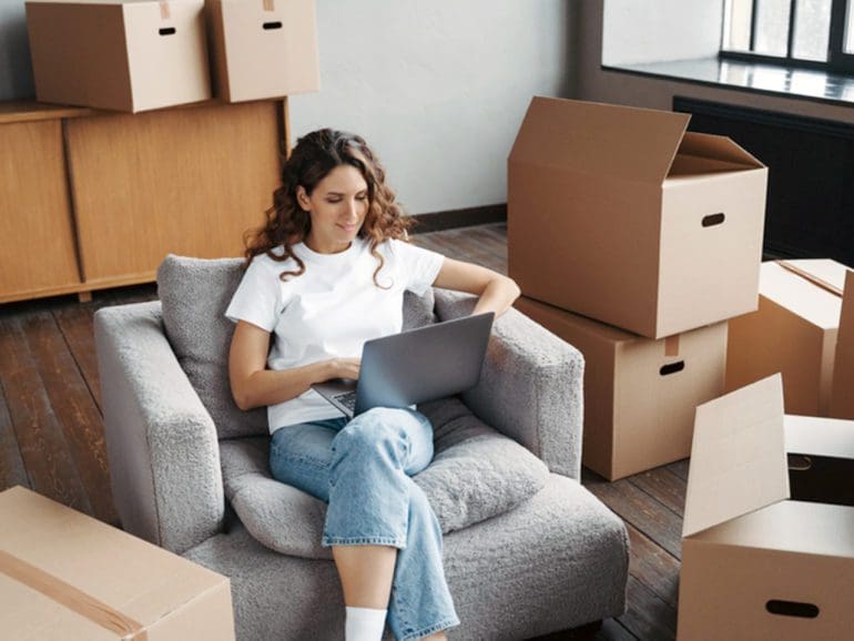 Happy young hispanic woman renting apartment online. Girl among cardboard boxes is using laptop and smiling. Happy homeowner is sitting in armchair with pc. Woman about to move.