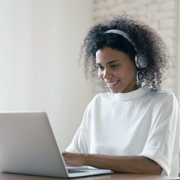 Smiling African American woman wearing headset using laptop in modern office, looking at screen and typing, listening to music, interpreter working online, learning computer course, lecture