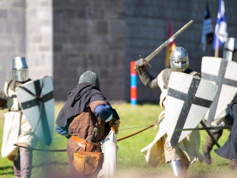 Located in Caboolture, the Abbey Medieval festival runs for a week and brings together lovers of all things old world. This image depicts several gentlemen dressed as knights in armor preparing retaliation to protect their king (and the castle you can see in the photo) during a battle in the LARP ring. The LARP battle take place in the large arena set upright in the heart of the festival. You can see the archer bracing to take his shot and the oncoming attack raising their shields in preparation. In this Image he is loading his bow with an arrow. This photo was taken at the festival on the 14th of July 2019 held at the Abbeystowe Museum.
